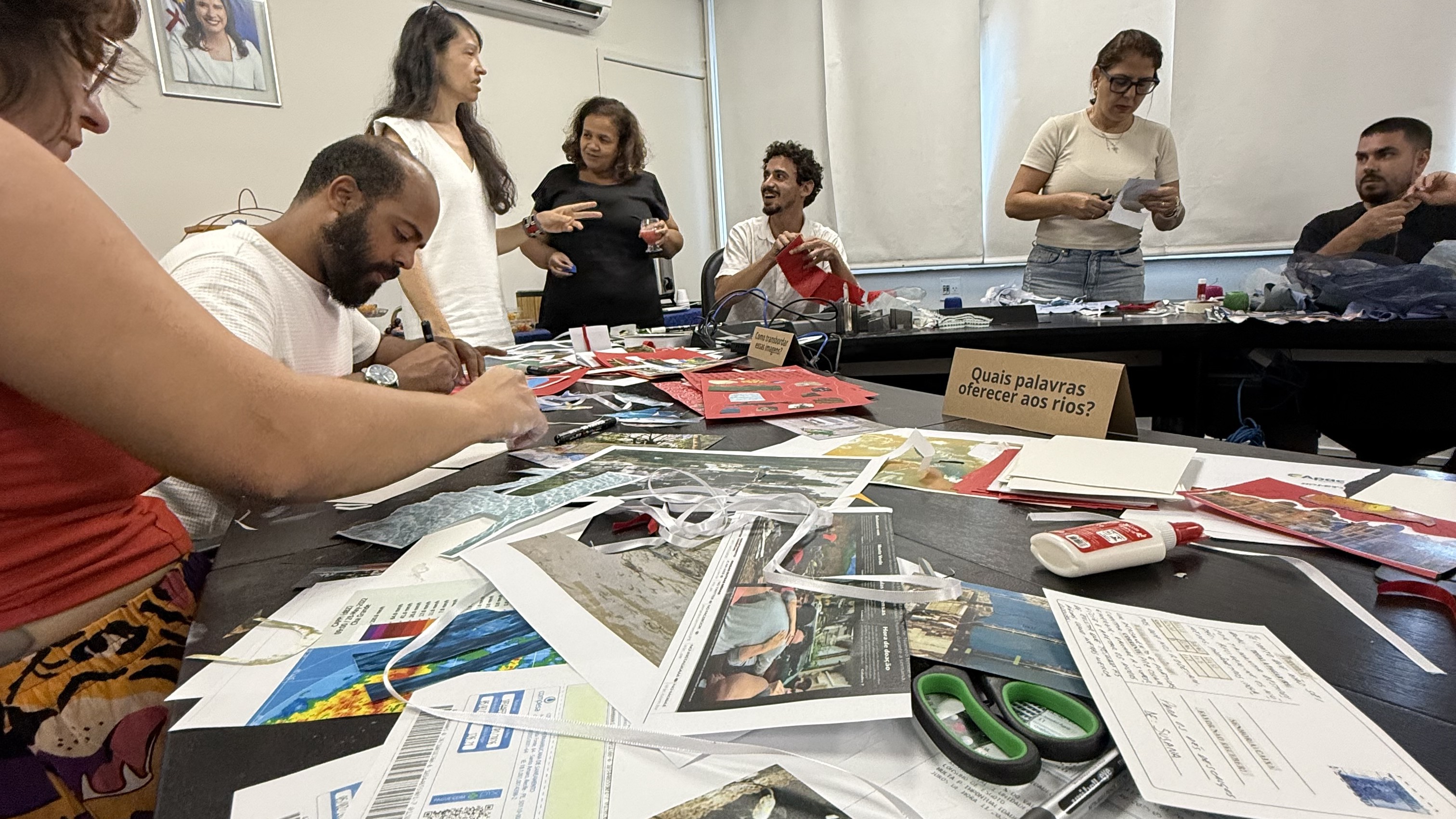Oficina coletiva com mesa cheia de materiais impressos, fotos e mapas — trabalho de comunicação e arte